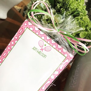 May include: Pink and green notepad with a tennis racket and ball design. The notepad has a pink and white diamond pattern border. The text "Jen Willson" is printed on the notepad.