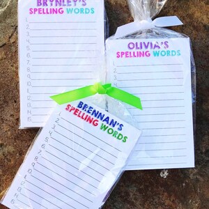 May include: Three white notepads with colorful text that reads "Brynley's Spelling Words", "Olivia's Spelling Words", and "Brennan's Spelling Words". Each notepad is wrapped in clear plastic and tied with a ribbon.