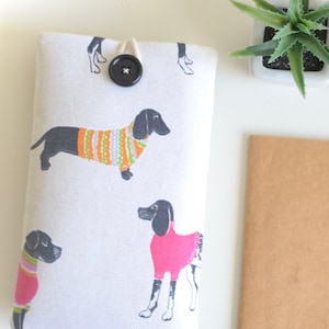May include: A white fabric phone case featuring a pattern of black dachshunds wearing colourful sweaters. The case has a button closure and a white string loop.