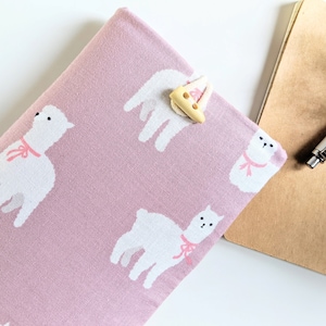 May include: A pink fabric book cover with a white alpaca pattern and a light brown button closure.