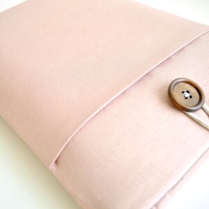 May include: A blush pink laptop sleeve with a flap closure and a wooden button. The sleeve has a white fabric trim around the edges.
