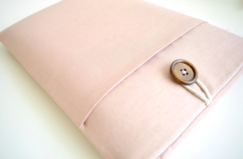 May include: A blush pink laptop sleeve with a flap closure and a wooden button. The sleeve has a white fabric trim around the edges.