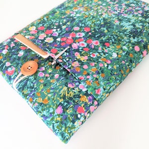 Pode incluir: Uma capa para laptop de tecido verde com um padrão floral com flores rosa, azuis e amarelas. A capa tem um fecho de botão de madeira e uma correia de couro castanho.