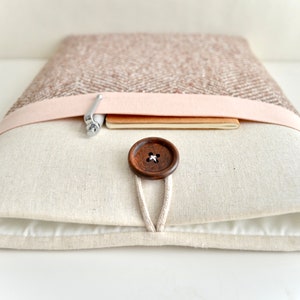 May include: A beige laptop sleeve with a brown button closure and a pink fabric flap. The sleeve has a pen and a notebook inside.