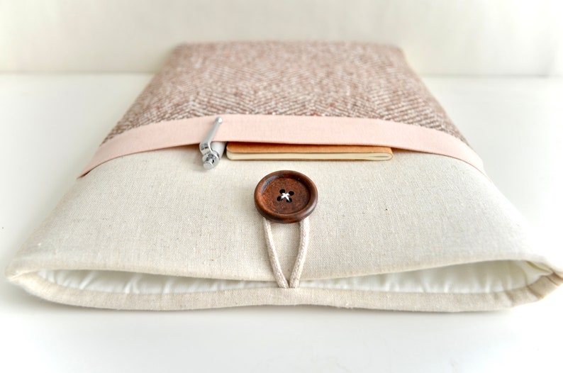 May include: A beige laptop sleeve with a brown button closure and a pink fabric flap. The sleeve has a pen and a notebook inside.