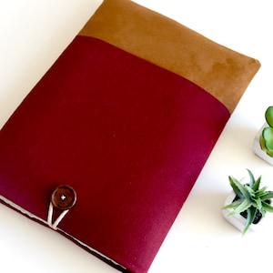 Può includere: Una custodia per laptop color bordeaux con una parte superiore in camoscio marrone e un solo bottone marrone. La custodia è su una superficie bianca.