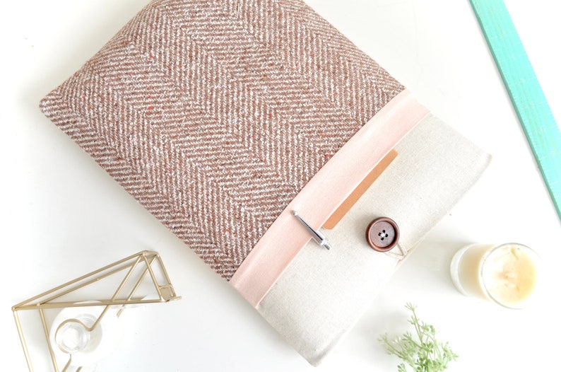 May include: A brown and white herringbone patterned laptop sleeve with a pink flap and a brown button. The flap has a brown leather-like pocket with a silver pen inside.