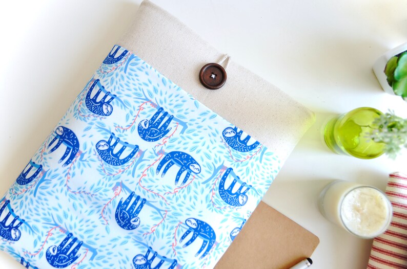 K&ouml;nnte beinhalten: Eine beige Laptoptasche mit einer Klappe und einem braunen Knopf. Die Tasche ist mit einem blauen und wei&szlig;en Stoff gef&uuml;ttert, der ein sich wiederholendes Muster von Faultieren zeigt, die an &Auml;sten h&auml;ngen.