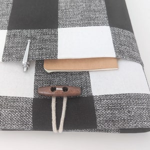 K&ouml;nnte beinhalten: Schwarze und wei&szlig;e karierte Laptop-H&uuml;lle aus Stoff mit einem braunen Holzknopf und einer wei&szlig;en Schnur. Die H&uuml;lle hat eine Tasche auf der Vorderseite mit einem silbernen Stift im Inneren.