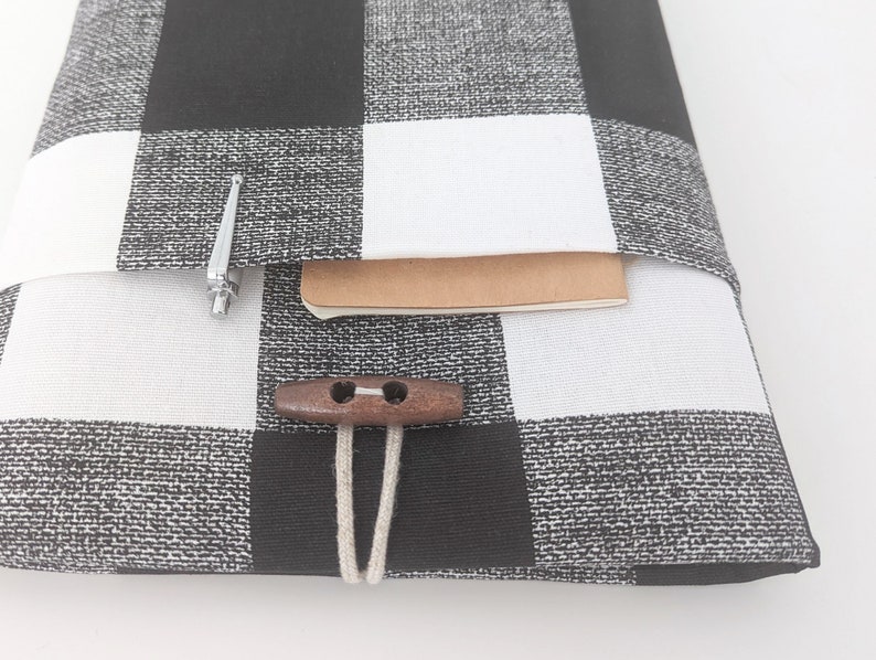 K&ouml;nnte beinhalten: Schwarze und wei&szlig;e karierte Laptop-H&uuml;lle aus Stoff mit einem braunen Holzknopf und einer wei&szlig;en Schnur. Die H&uuml;lle hat eine Tasche auf der Vorderseite mit einem silbernen Stift im Inneren.