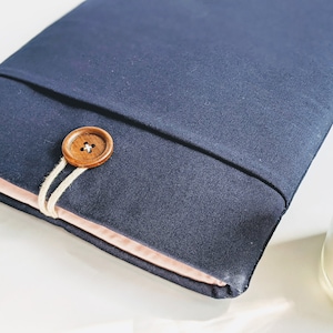 Op de afbeelding: Een marineblauwe stoffen hoes met een lichtroze binnenkant, vastgezet met een houten knoop en touwsluiting. De hoes is ontworpen om een tablet of e-reader in te bewaren. De knoop is rond en heeft een natuurlijke houtnerf.