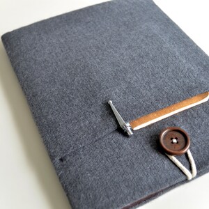 Peut inclure: Une housse pour ordinateur portable en tissu gris avec un bouton marron et une petite boucle en m&eacute;tal. La housse a une poche sur le devant avec un rabat en cuir marron.