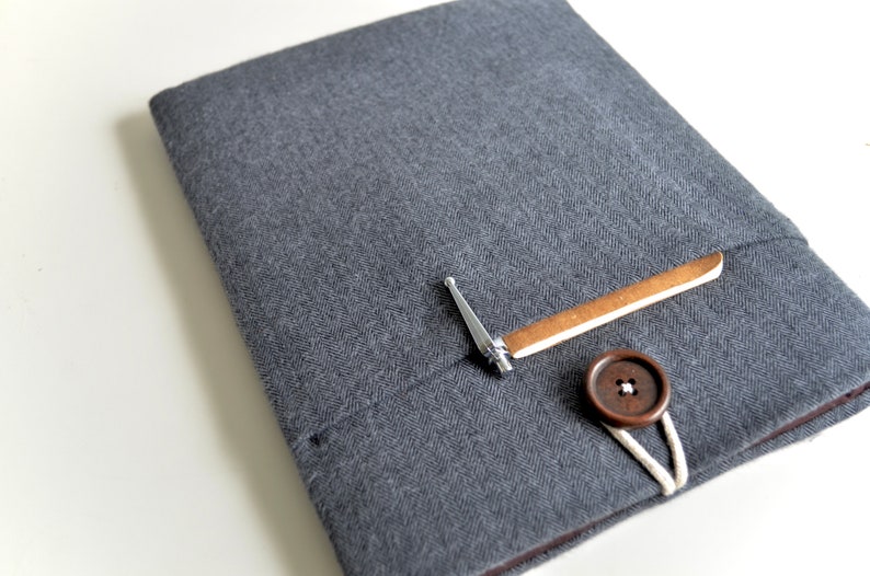 Peut inclure: Une housse pour ordinateur portable en tissu gris avec un bouton marron et une petite boucle en m&eacute;tal. La housse a une poche sur le devant avec un rabat en cuir marron.
