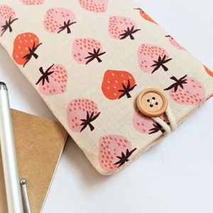 May include: A beige fabric case with a repeating pattern of pink and red strawberries. The case is secured with a wooden button and a white cord loop. A silver stylus and a brown notebook are in the background.