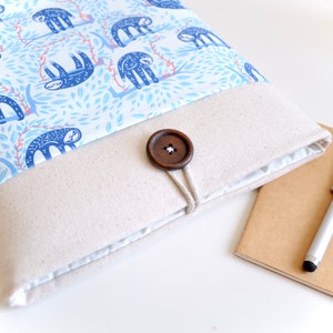 K&ouml;nnte beinhalten: Eine beige Laptop-H&uuml;lle aus Canvas mit einer blauen und wei&szlig;en Stoffklappe mit Faultier-Motiv. Die Klappe wird mit einem braunen Holzknopf und einer Schnur befestigt.