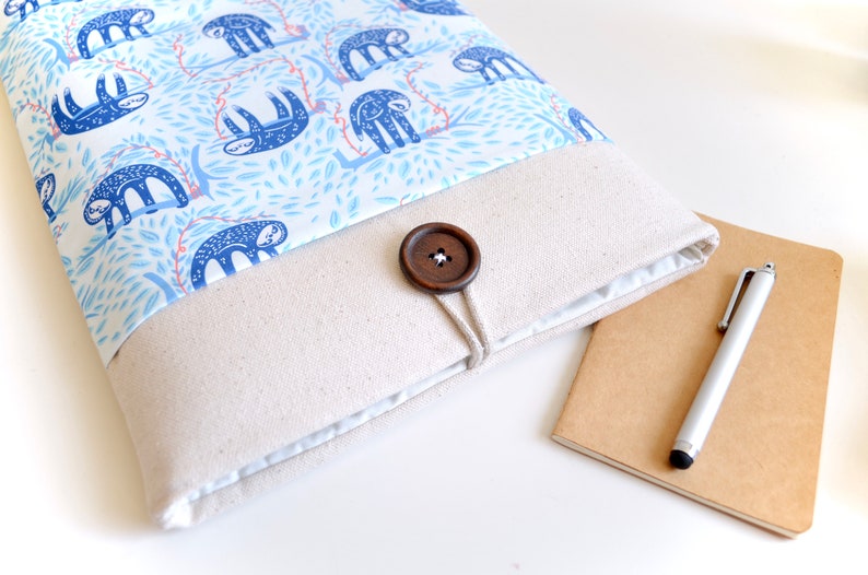 K&ouml;nnte beinhalten: Eine beige Laptop-H&uuml;lle aus Canvas mit einer blauen und wei&szlig;en Stoffklappe mit Faultier-Motiv. Die Klappe wird mit einem braunen Holzknopf und einer Schnur befestigt.