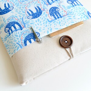 K&ouml;nnte beinhalten: Eine wei&szlig;e Laptop-H&uuml;lle aus Canvas mit einem blauen und wei&szlig;en Sloth-Muster als Futter. Die H&uuml;lle hat einen braunen Knopfverschluss und eine kleine Tasche auf der Vorderseite.
