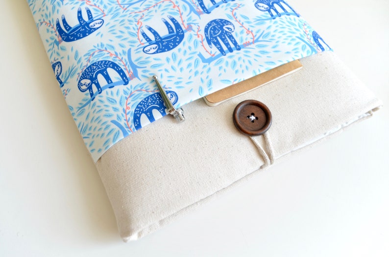 K&ouml;nnte beinhalten: Eine wei&szlig;e Laptop-H&uuml;lle aus Canvas mit einem blauen und wei&szlig;en Sloth-Muster als Futter. Die H&uuml;lle hat einen braunen Knopfverschluss und eine kleine Tasche auf der Vorderseite.