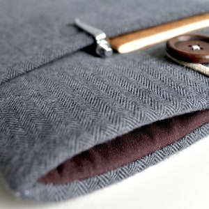 Peut inclure: Une housse pour ordinateur portable en tissu gris &agrave; chevrons avec une doublure en tissu brun. La housse a un rabat avec une fermeture en m&eacute;tal et un bouton brun avec un cordon blanc.
