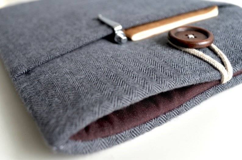 Peut inclure: Une housse pour ordinateur portable en tissu gris &agrave; chevrons avec une doublure en tissu brun. La housse a un rabat avec une fermeture en m&eacute;tal et un bouton brun avec un cordon blanc.