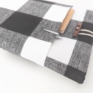 K&ouml;nnte beinhalten: Ein Notizbuchcover aus schwarzem und wei&szlig;em kariertem Stoff mit braunem Knopfverschluss und einem Stift, der in die Tasche gesteckt ist.
