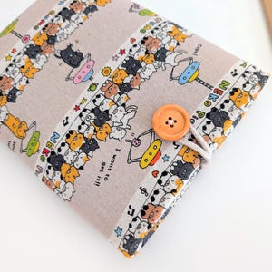 May include: A beige fabric book cover featuring a repeating pattern of cartoon cats in various colors. The cover has an orange button closure and the text "NEKO Pic" and "I want to get it!!".