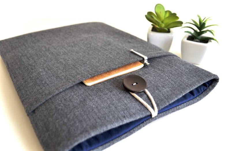 May include: A gray herringbone fabric laptop sleeve with a blue lining, a brown notebook inside, and a black button closure.