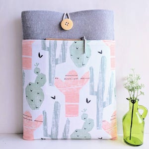 Puede incluir: Una funda para portátil gris y blanca con estampado de cactus. La funda tiene una solapa con cierre de botón.