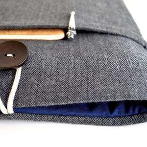 Puede incluir: Una funda para portátil de tela gris de espiga con forro azul y cierre de botón marrón.