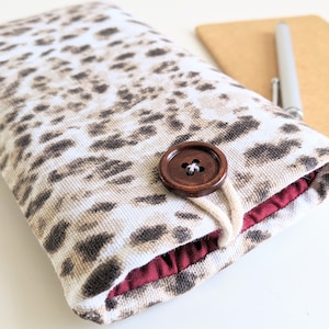 May include: A white and brown leopard print fabric phone case with a brown button closure and a maroon lining.