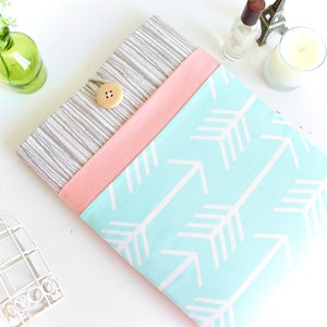 May include: A light blue laptop sleeve with a white arrow pattern. The sleeve has a pink fabric flap and a gray fabric backing with a wooden button.