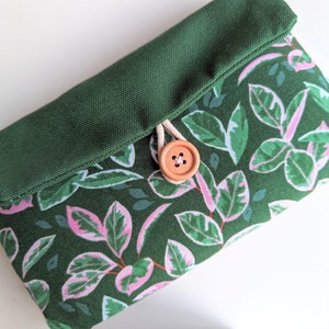 May include: A green canvas pouch with a flap closure and a wooden button. The pouch has a pink and green leaf pattern.