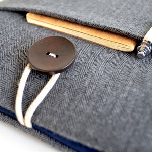 May include: A gray herringbone fabric laptop sleeve with a brown button closure and a white cord. A brown notebook and a silver pen are tucked inside the sleeve.