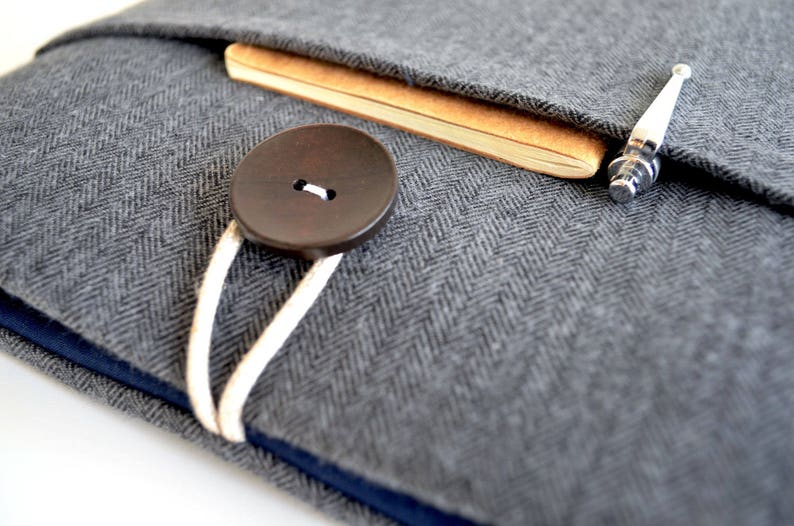 May include: A gray herringbone fabric laptop sleeve with a brown button closure and a white cord. A brown notebook and a silver pen are tucked inside the sleeve.