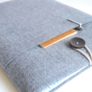 May include: A gray herringbone fabric laptop sleeve with a brown leather flap and a black button closure.