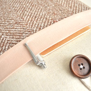 May include: A brown and white tweed fabric laptop sleeve with a pink velvet trim, a silver pen, and a brown wooden button.