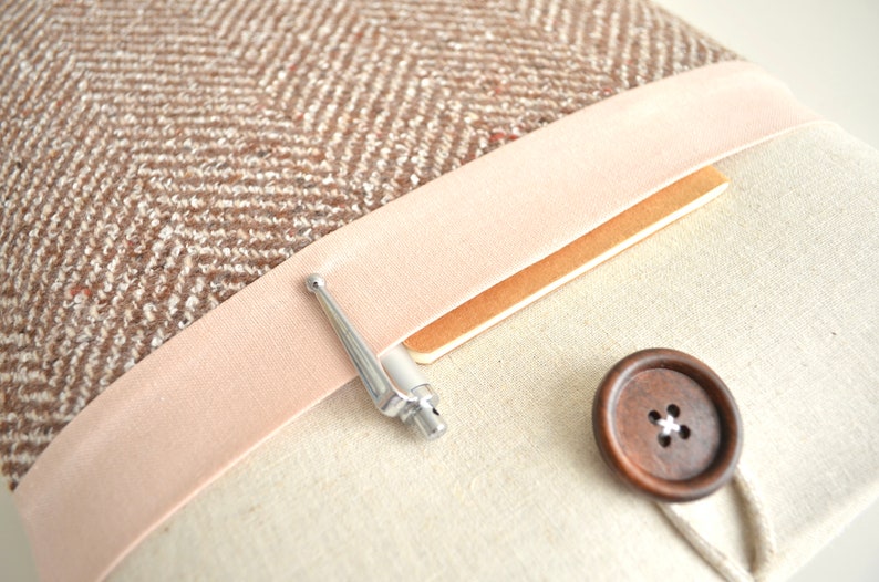 May include: A brown and white tweed fabric laptop sleeve with a pink velvet trim, a silver pen, and a brown wooden button.