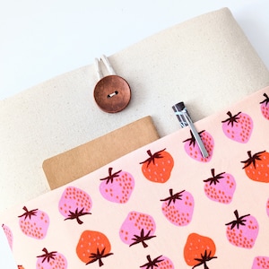 May include: A beige canvas laptop sleeve with a pink and orange strawberry print fabric lining. The sleeve has a brown wooden button and a loop for a pen.