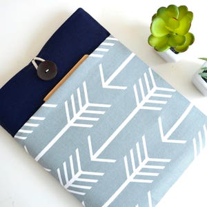 May include: A gray and navy blue laptop sleeve with a white arrow pattern. The sleeve has a button closure and a flap.