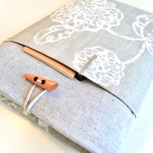 May include: A gray linen notebook cover with a white floral design. The cover has a flap that folds over and closes with a wooden button. A small pocket on the front of the cover holds a notebook.