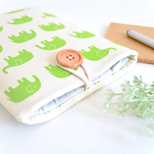 Puede incluir: Una funda de tela color crema con un patrón repetido de elefantes verdes. La funda se asegura con un botón de madera y un cierre de cuerda. Un cuaderno, un bolígrafo y una ramita de vegetación están en el fondo.