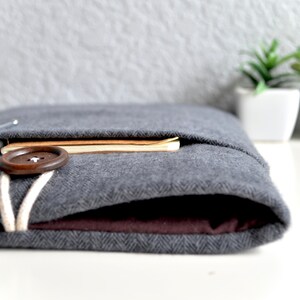 Peut inclure: Une pochette pour ordinateur portable en feutre gris avec un bouton marron et un cordon blanc. La pochette est pli&eacute;e et un carnet est visible &agrave; l'int&eacute;rieur.