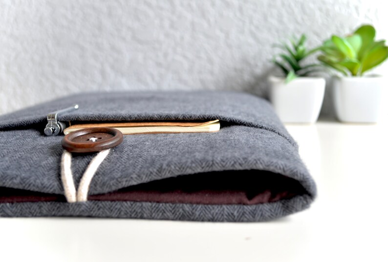 Peut inclure: Une pochette pour ordinateur portable en feutre gris avec un bouton marron et un cordon blanc. La pochette est pli&eacute;e et un carnet est visible &agrave; l'int&eacute;rieur.