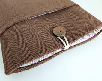 Puede incluir: Una funda para portátil de color marrón con un patrón de espiga en marrón oscuro. La funda se cierra con un botón redondo de madera y un cordón elástico blanco. El forro interior es visible, mostrando una tela clara con estampado floral.