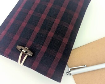 May include: A red and black plaid notebook cover with a wooden toggle closure. The cover is made of a soft, textured fabric.