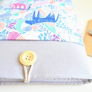 K&ouml;nnte beinhalten: Eine graue Laptoptasche mit einer wei&szlig;en Stoffklappe, die mit einem bunten Muster aus Bl&auml;ttern, Tieren und geometrischen Formen verziert ist. Die Klappe wird mit einem Holzknopf und einer Schnur geschlossen.