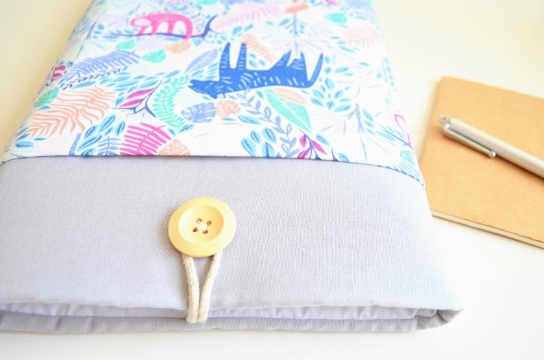 K&ouml;nnte beinhalten: Eine graue Laptoptasche mit einer wei&szlig;en Stoffklappe, die mit einem bunten Muster aus Bl&auml;ttern, Tieren und geometrischen Formen verziert ist. Die Klappe wird mit einem Holzknopf und einer Schnur geschlossen.