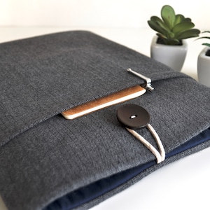May include: A grey herringbone laptop sleeve with a zipper closure and a brown leather button. The sleeve has a pocket on the front for a notebook or other small items.