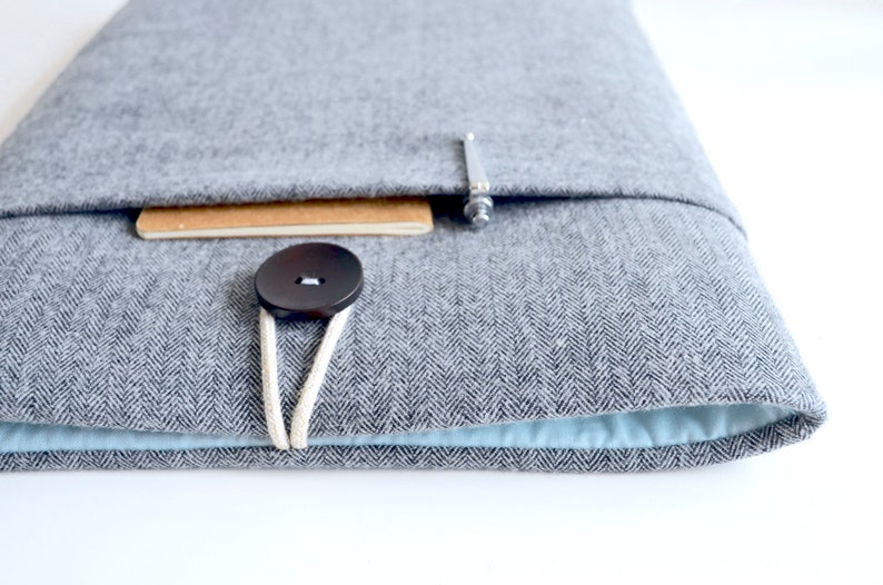 Puede incluir: Una funda gris de fieltro para port&aacute;til con un patr&oacute;n de espiga. La funda tiene un cierre de bot&oacute;n con un cord&oacute;n de color claro y un peque&ntilde;o cuaderno asomando. El interior es de color azul claro.