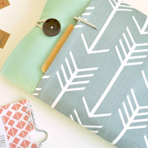 Puede incluir: Una funda para portátil de tela gris y blanca con un patrón de flechas repetido. La funda tiene una solapa verde menta con un botón negro y un clip metálico plateado.